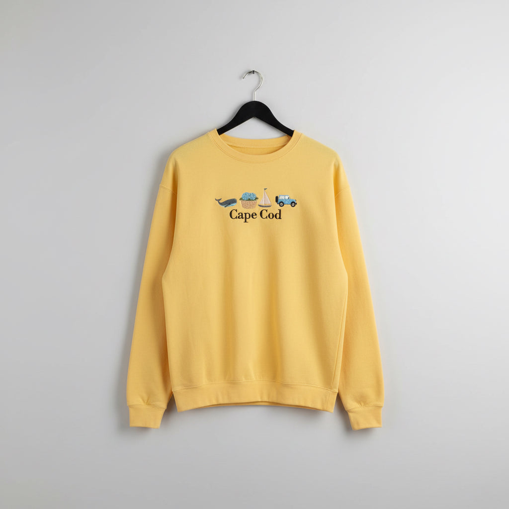 A yellow long-sleeve t-shirt with a 'Cape Cod' whale, clam, sailboat, and car graphic on the chest, hanging outdoors on a sunny day.