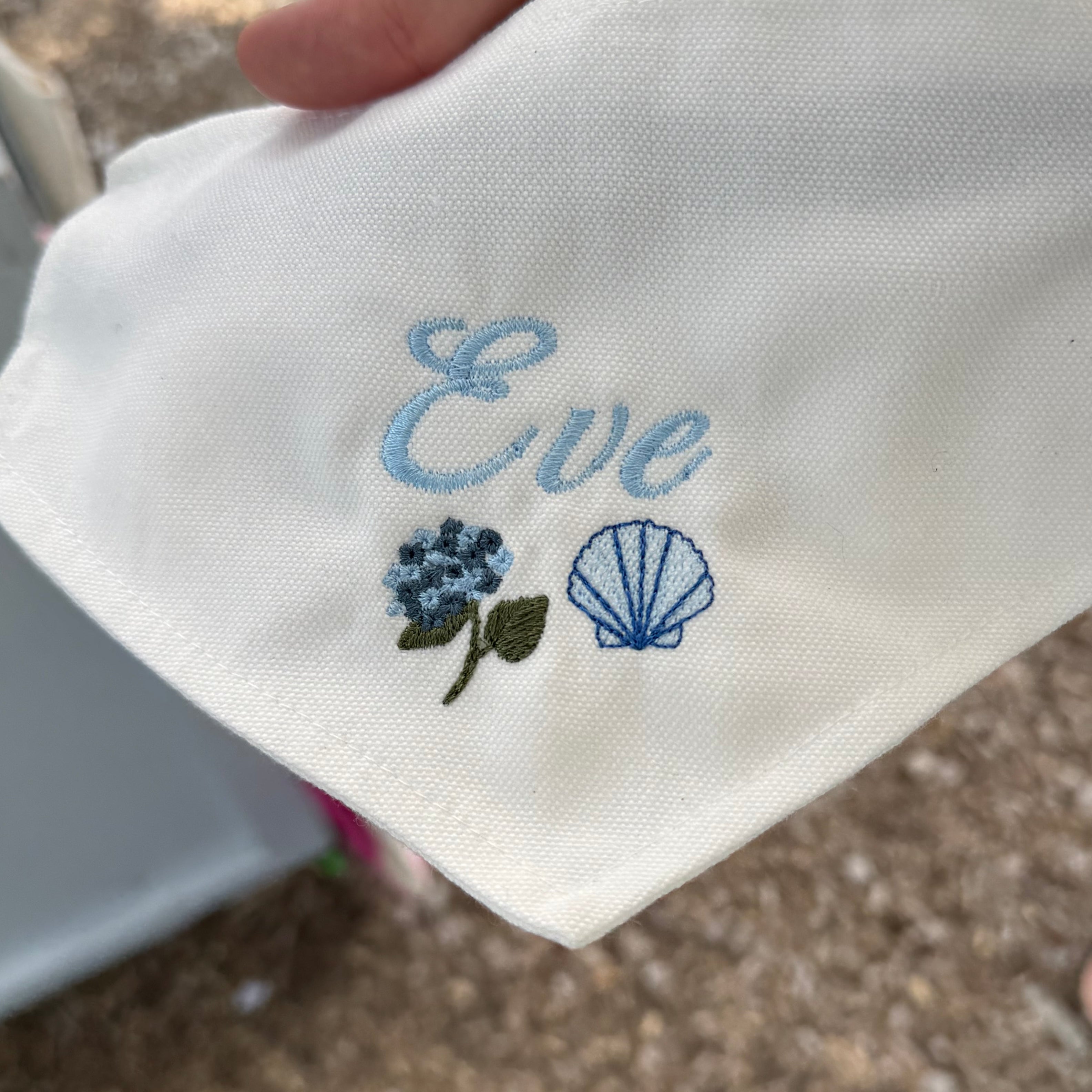 A close-up of a white fabric bandana or pocket square held by a hand, featuring light blue embroidered text reading 'Eve' and small floral and shell motifs in blue, green, and white. The background shows an outdoor setting with gravel and other people partially visible.