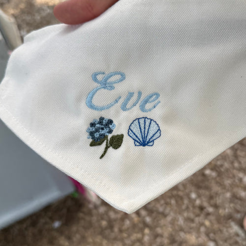 A close-up of a white fabric bandana or pocket square held by a hand, featuring light blue embroidered text reading 'Eve' and small floral and shell motifs in blue, green, and white. The background shows an outdoor setting with gravel and other people partially visible.