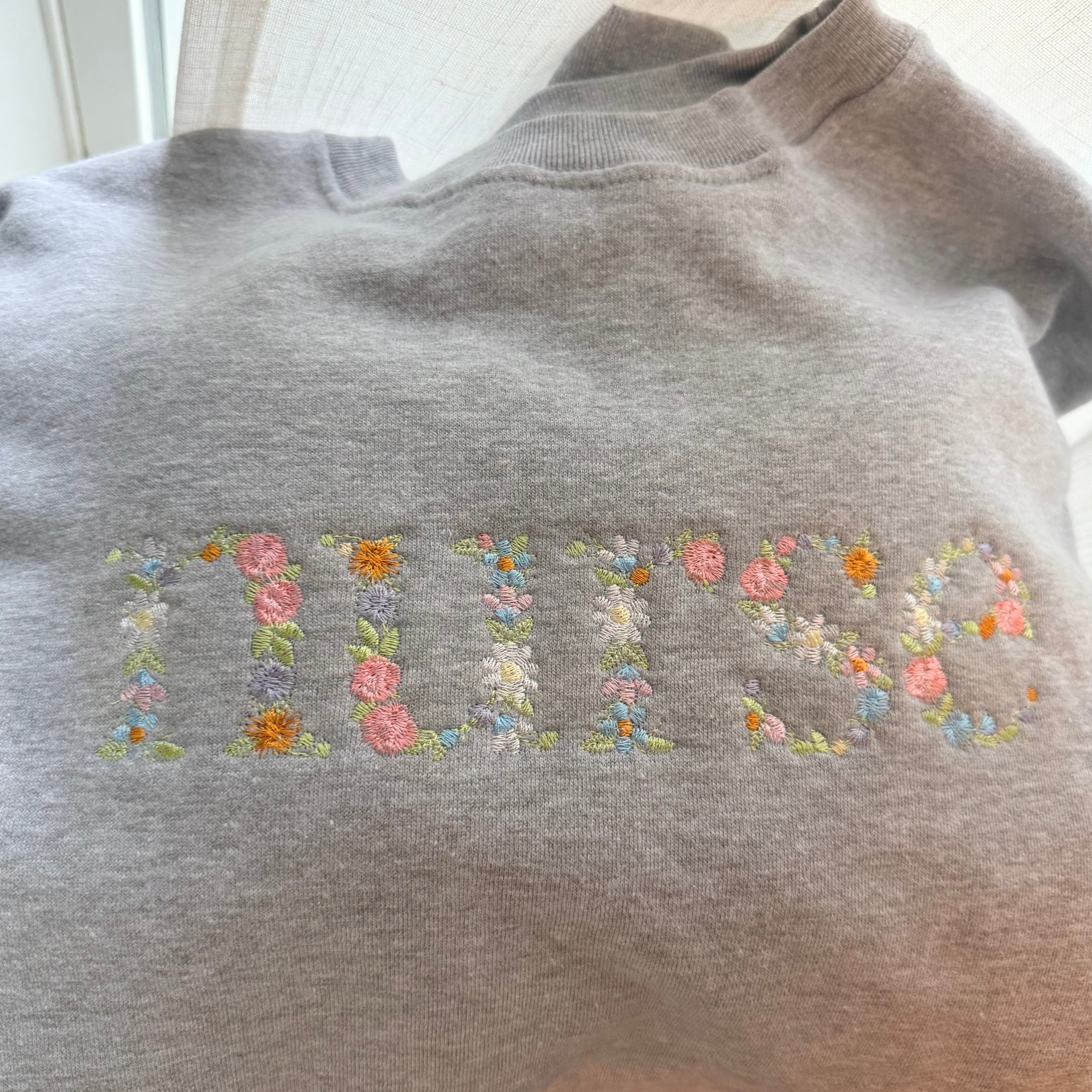 A light gray crewneck sweatshirt featuring the word 'NURSE' embroidered across the chest in a floral pattern with pink, orange, yellow, and white flowers and green leaves, shown in a casual indoor setting.