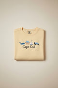 A pale yellow t-shirt with embroidered nautical-themed graphics and the text 'Cape Cod' in black script. The embroidery includes a blue car, sailboat, ice cream cone, blue whale, and a small pink boat.