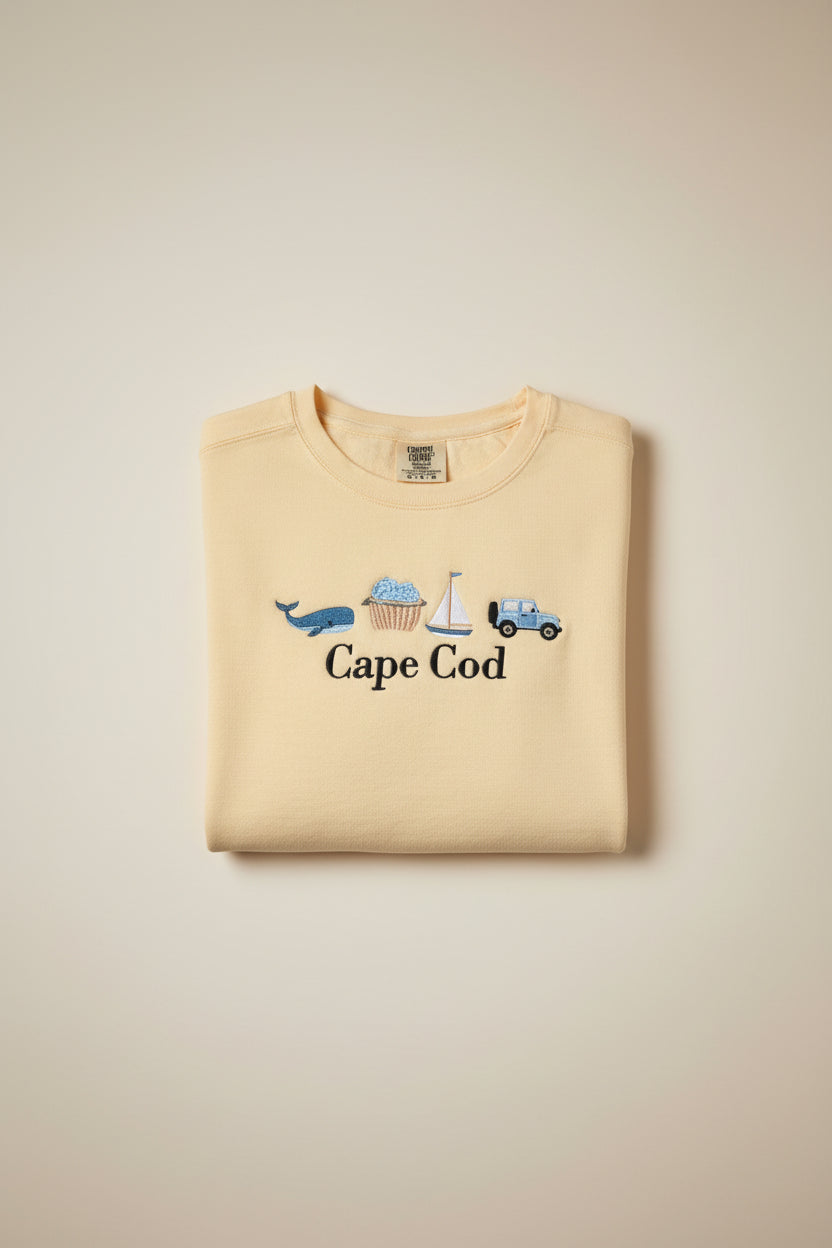 A pale yellow t-shirt with embroidered nautical-themed graphics and the text 'Cape Cod' in black script. The embroidery includes a blue car, sailboat, ice cream cone, blue whale, and a small pink boat.