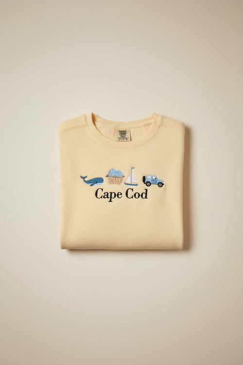 A pale yellow t-shirt with embroidered nautical-themed graphics and the text 'Cape Cod' in black script. The embroidery includes a blue car, sailboat, ice cream cone, blue whale, and a small pink boat.