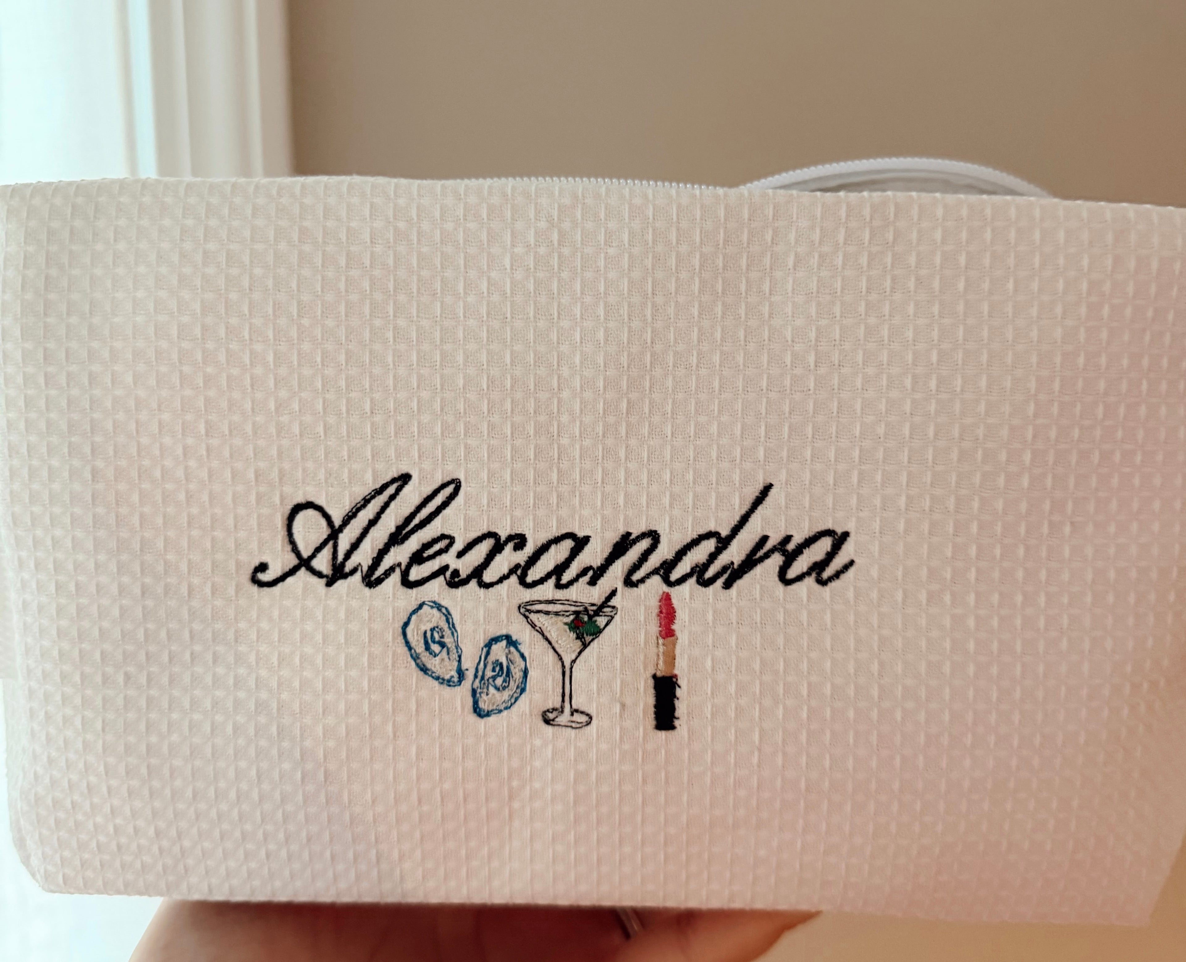 Beige pouch with embroidered name and icons held by a hand